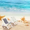 Two miniature deck chairs and seashells on a sandy beach with the ocean in the background. Produktbild-Vorschau