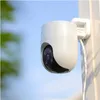 A white security camera mounted on a white pole against a blue sky with blurred green foliage on the left. Produktbild-Vorschau