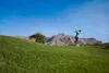 A golfer swings a club on a golf course with mountains in the background. Produktbild-Vorschau