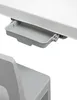 A close-up view of a gray plastic tray attached to the underside of a white surface, with a portion of a gray chair visible below. Náhled obrázku produktu