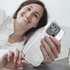 A woman is lying in bed, smiling and holding a transparent phone case with a Redmi phone inside. Produktbild-Vorschau