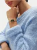 A person wearing a light blue fuzzy sweater and a gold watch with a beige strap. Produktbild-Vorschau