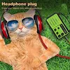An orange cat wearing headphones and sunglasses is lying on grass, with a green radio next to it. Produktbild-Vorschau