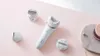 A white electric epilator with multiple attachments is displayed on a pink surface, along with some decorative items. Náhľad obrázku produktu