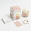 A product shot of a cute chick-shaped night light, its packaging, and accessories on a white background. Produktbild-Vorschau