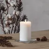 A white LED candle with a flickering flame sits on a square glass coaster on a wooden table. In the background, there is a decorative arrangement of cotton bolls and dried branches. Podgląd zdjęcia produktu