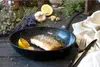 A cooked salmon fillet in a frying pan, with lemon slices and herbs, surrounded by ingredients like garlic and lavender. Anteprima dell’immagine prodotto