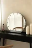 A round makeup mirror with lights sits on a black vanity table, reflecting a chair and curtains. Two vases and a makeup bottle are also on the table. Produktbild-Vorschau