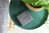 A gray Fujifilm Instax Link Wide printer is placed on a green round table next to a potted plant. Produktbild-Vorschau