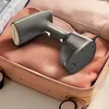 A handheld garment steamer is placed inside an open suitcase, resting on folded clothing. Náhľad obrázku produktu