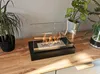 A bio-fireplace is placed on a wooden surface, with plants in the background. Produktbild-Vorschau