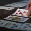 A hand is holding a playing card over a table with other cards spread out. Produktbild-Vorschau