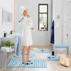 A woman in a white bathrobe and towel is standing in a modern bathroom, surrounded by blue and white striped bath mats and towels. Produktbild-Vorschau