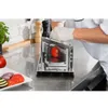 A chef in a white uniform and gloves is using a stainless steel tomato slicer in a professional kitchen setting. Sliced vegetables are on a plate in the foreground. Voir les images du produit