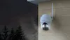A white security camera mounted on a wooden wall at night, with trees visible in the dark background. Produktbild-Vorschau