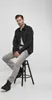 A man is sitting on a stool, wearing a black jacket and gray jeans. Produktbild-Vorschau
