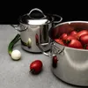 Two stainless steel pots, one filled with tomatoes, and a single onion with green stems are arranged on a gray surface against a black background. Produktbild-Vorschau