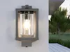 A modern outdoor wall lamp mounted on a white wall, with a blurred background showing a garden and a deck. Anteprima dell’immagine prodotto