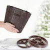A person is holding a brown Christmas tree stand, with two other stands on a marble surface in the foreground. A blurred Christmas tree is in the background. Voir les images du produit