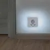 A white Hama LED night light with a power socket and two USB ports is mounted on a light-colored wall near the floor. The night light is emitting a soft white glow. A wooden skirting board runs along the bottom of the wall, and a wooden floor is visible. Voir les images du produit