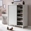 A white shoe cabinet with sliding doors, filled with shoes, is placed in a hallway next to stairs. A tote bag and two heels are on the floor in front of it. Náhled obrázku produktu