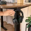 A hand is shown hanging a pair of black and green headphones on a black stand attached to a brown desk. Produktbild-Vorschau