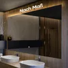 A modern bathroom with a large mirror reflecting a shower and two sinks. The mirror has illuminated text and is mounted above a black cabinet. Produktbild-Vorschau