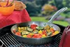 A frying pan filled with colorful vegetables is placed on a grill, with a table covered in a red tablecloth and a bowl of chili peppers in the background. Voir les images du produit