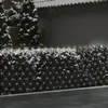 A close-up view of a lit-up Christmas light net draped over a dark hedge, with snow on top and trees in the background. Produktbild-Vorschau