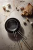 A top-down view of a black frying pan with a smaller pan inside, placed on a marble-like surface with scattered ingredients like onions, garlic, and ginger. Produktbild-Vorschau