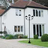A black metal street lamp with three lights stands in front of a white house with a red roof. Produktbild-Vorschau
