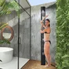 A woman is showering outdoors next to a concrete wall and lush greenery. Produktbild-Vorschau
