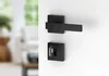 A black door handle and key plate are shown against a white background. The background is blurred and shows a hint of a room interior with furniture and a window. Produktbild-Vorschau