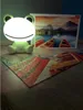 A frog-shaped LED lamp sits on a wooden surface next to a partially completed jigsaw puzzle and its box, which depicts a scenic mountain lake with boats. Náhľad obrázku produktu