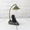 A decorative lamp ornament shaped like a faceless man sitting on a log, with a flexible gooseneck lamp arm and a woven lampshade. Produktbild-Vorschau