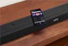 A black JBL soundbar with a smartphone placed on top, displaying a music app interface, resting on a wooden surface. Produktbild-Vorschau