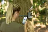 A woman is checking her phone while standing in front of a trail camera mounted on a tree. Náhled obrázku produktu