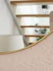 A close-up view of a round mirror with a gold rim, set against a textured pink surface. The background shows blurred wooden stairs and a white lampshade. Produktbild-Vorschau
