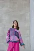 A woman stands in front of a concrete wall wearing a colorful sweater and pink pants. Produktbild-Vorschau