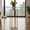 A plant stand with a plant on it, placed in a room with large windows and a view of trees. Produktbild-Vorschau