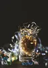 A glass jar filled with a pinecone and fairy lights, surrounded by decorative glass marbles and another pinecone on a wooden surface. Náhľad obrázku produktu