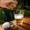 A hand is holding a wooden smoking device over a glass of whiskey, with a small tin of wood chips next to it. Voir les images du produit