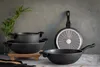 A set of black cookware, including pots and pans, arranged on a surface with a textured gray background and a vase of greenery. Náhľad obrázku produktu