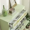 A close-up view of a light green shoe cabinet with an open drawer, displaying watches and stationery. The top surface holds a green vase and a small wooden radio on a book. Shoes are visible in the lower compartments. Voir les images du produit