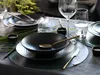 A table setting with black stoneware plates, bowls, cutlery, and glassware, decorated with green grass. Produktbild-Vorschau