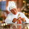 A chef in a white uniform and hat is holding a chocolate Santa Claus figure. There are Christmas ornaments and greenery on the table in front of him. The background is blurred and appears to be a festive indoor setting. Náhled obrázku produktu
