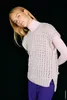 A woman wearing a knitted vest over a turtleneck and pants, posing against a black background. Produktbild-Vorschau