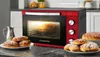 A red and black Kalorik mini oven is shown with a cake baking inside. Several pastries are arranged on plates in the foreground, and a metal pitcher is to the left. Produktbild-Vorschau