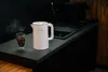 A white electric kettle is shown on a dark countertop next to a glass of coffee, with steam rising from the kettle. Produktbild-Vorschau
