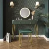 A dark green vanity table with gold legs and a matching stool, set against a dark green and gold geometric wallpaper. A round mirror with LED lighting is above the table, and a shopping bag is on the floor. Náhľad obrázku produktu